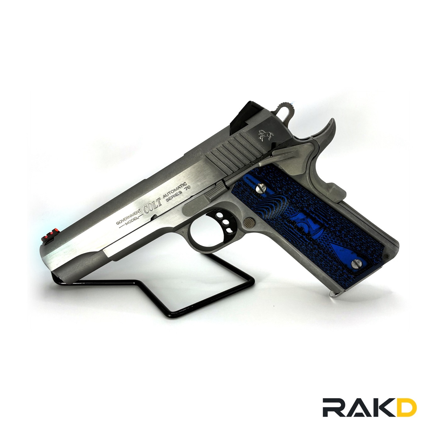 A stainless Colt Government Model Series 70 pistol on a wire stand, three-quarter view, with blue and black G10 grips and a red fiber-optic front sight.